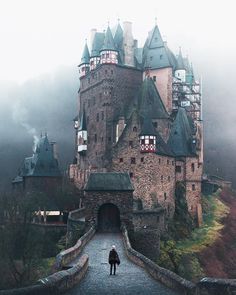 a person standing on a bridge in front of a castle with towers and turrets, surrounded by fog