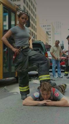 two fire fighters laying on the ground in front of a bus and people standing around