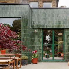 an outdoor patio with chairs, tables and potted plants in front of a green tiled building