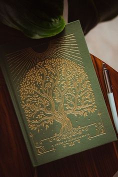 a green book sitting on top of a wooden table next to a pen and plant