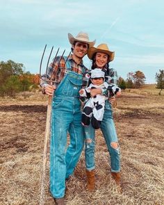 a man and woman in overalls holding a baby while standing on dry grass with pitchforks