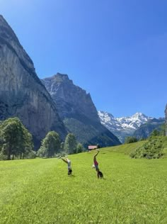 two people are playing with a dog in a field near mountains and snow capped peaks