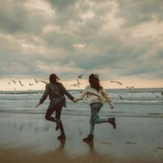 two people running on the beach with seagulls flying in the sky behind them