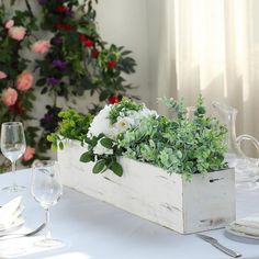 an arrangement of flowers and greenery in a wooden box on a table with wine glasses