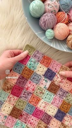 two hands are holding a crocheted square in front of some balls of yarn