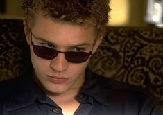 a young man wearing sunglasses sitting in a chair looking down at his cell phone while he is staring into the camera