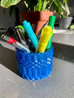 a blue cup filled with pens and markers on top of a table next to a potted plant