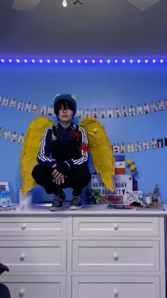 a young boy sitting on top of a dresser in front of a blue wall with angel wings