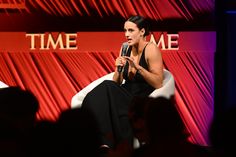 a woman sitting in a chair talking on a stage with a microphone to her left