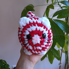 a hand holding up a small crocheted red and white kitty ball with black eyes