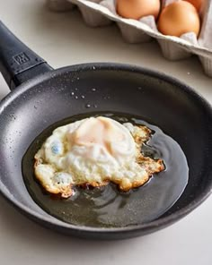 an egg frying in a skillet on a counter next to eggs and a carton of eggs