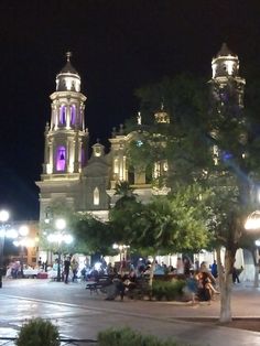 people are sitting on benches in front of a large building at night with street lights