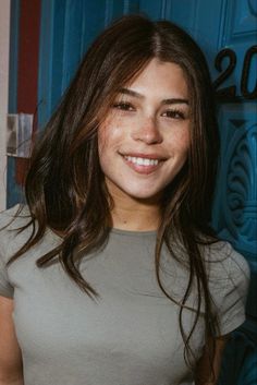 a woman with long brown hair smiling at the camera and wearing a gray shirt in front of a blue door