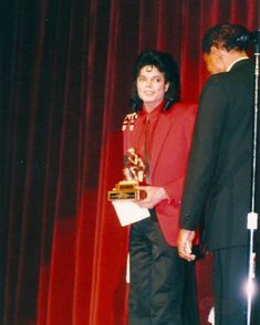 a man standing next to a woman in front of a red curtain holding an award