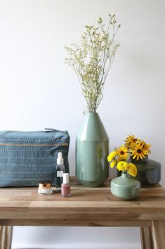 a wooden table topped with a green vase filled with yellow flowers next to a blue bag