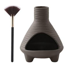 a grey vase with a black handle next to a large brush on a white background