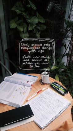 an open book on a table with notebooks and pens next to it, near a plant