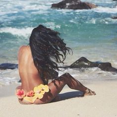 a woman sitting on top of a sandy beach next to the ocean with flowers in her hair