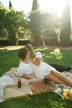 a man and woman sitting on a blanket in the grass with pizza, beer bottles and flowers