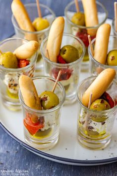 small cups filled with olives, pickles and bread sticks on a white plate