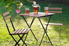 two chairs and a table with drinks on it in the grass near a flower bush