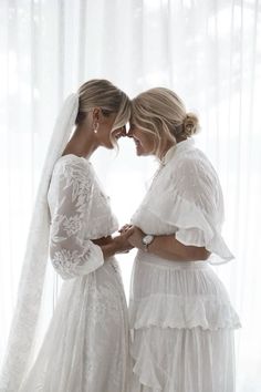 two women standing next to each other in front of a window wearing white dresses and veils