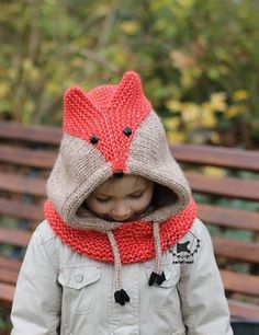 a little boy wearing a knitted fox hat and scarf in front of a park bench