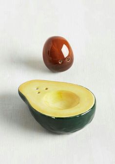 an avocado shaped bowl next to a red apple on a white surface, with one cut in half