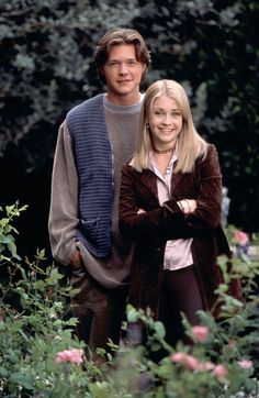 a man and woman standing next to each other in front of bushes with pink flowers