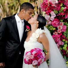 a bride and groom kissing each other in front of a flower covered wall with pink flowers