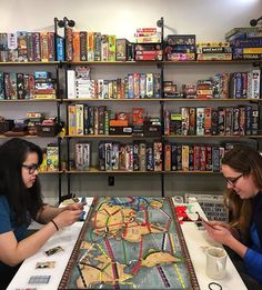 two women sitting at a table looking at their cell phones in front of a board game