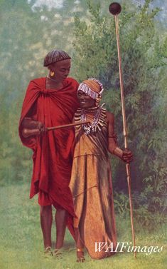 two african women standing next to each other in front of trees and bushes, one holding a stick