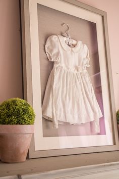 a baby's white dress hangs on the wall next to a potted plant