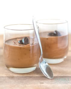 two glasses filled with dessert sitting on top of a wooden table next to a spoon