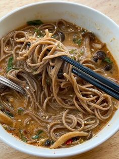 a white bowl filled with noodles and vegetables on top of a wooden table next to chopsticks