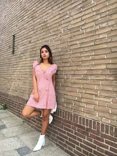 a woman leaning against a brick wall wearing a pink dress and white tennis shoes with her legs crossed