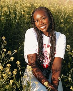 a woman with long braids sitting in the middle of a field smiling at the camera