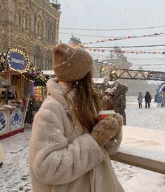 a woman standing in the snow holding a cup of coffee and looking at christmas lights