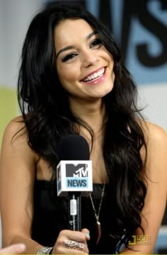 a smiling woman holding a microphone in front of a tv studio sign with the news logo on it