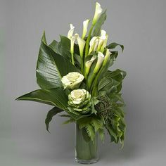 a vase filled with white flowers and greenery on top of a gray surface next to a plant