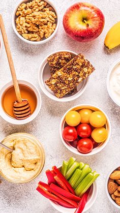 various fruits and vegetables are arranged in bowls on a white counter top, including bananas, apples, carrots, nuts, honey