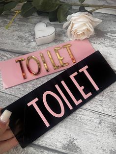 a pink and black sign that says toilet next to a white rose on a wooden table