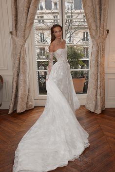 a woman in a white wedding dress standing by a window with her hands on her hips