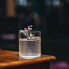 a glass filled with water sitting on top of a wooden table next to a purple flower