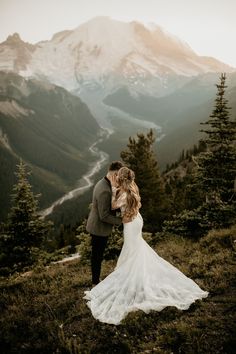 a bride and groom standing on top of a mountain looking at each other with mountains in the background