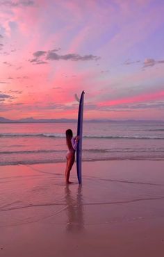 a woman holding a surfboard on top of a beach next to the ocean at sunset