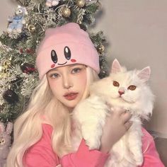 a woman holding a white cat in front of a christmas tree wearing a pink hat