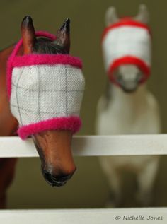 two horses wearing sweaters on their heads behind a white fence and one horse is looking at the camera