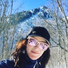 a woman wearing sunglasses and a hat taking a selfie in front of some trees