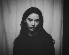 a black and white photo of a woman with long hair looking at the camera while standing in front of a curtain